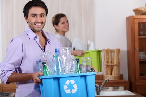 A young man with dark hair and a beard, wearing a light purple shirt, is smiling and holding a blue recycling bin filled with mostly green and transparent glass bottles. Behind him, a woman with brown hair tied back, dressed in a light top, stands in a domestic kitchen setting, next to a green plastic container and a wooden cabinet. The background includes a kitchen countertop, a sink, and wooden shelving units, suggesting an indoor environment related to household waste collection or rubbish clearance. The scene appears well-lit with natural or bright indoor lighting, emphasizing the focus on waste sorting and removal activities. This image visually supports rubbish removal services provided by Hillingdon House Clearance, highlighting environmentally conscious waste disposal practices in a suburban or residential area.