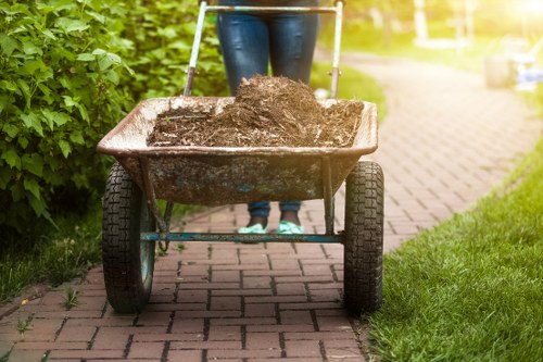A person pushing a metal wheelbarrow filled with dark, loose soil or compost along a paved pathway adjacent to a grassy garden area. The wheelbarrow has black rubber tires and metal handles, with soil visibly piled high and slightly spilling over the sides. The individual is wearing dark jeans and a long-sleeved top, though their upper body is not visible in the image. The environment includes green bushes on the left side of the pathway, with sunlight illuminating the scene and casting a warm glow. This scene depicts a gardening or clearance activity, relevant to rubbish removal and garden waste collection services offered by Hillingdon House Clearance, situated in the Hillingdon area.